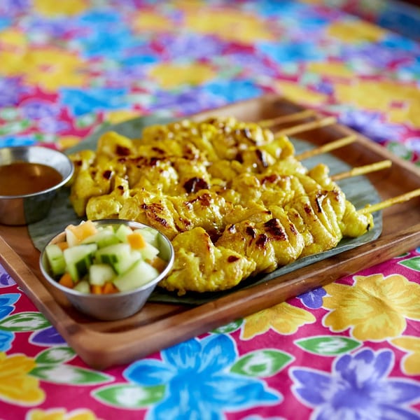 Chicken satay skewers with peanut sauce and cucumber salad on a colorful floral tablecloth.