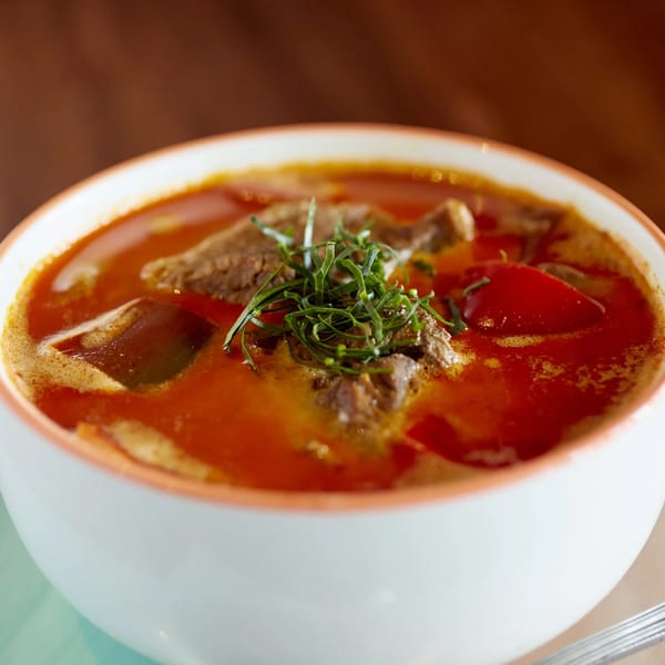 Close-up of a vibrant red curry soup with beef and red peppers, garnished with fresh herbs.